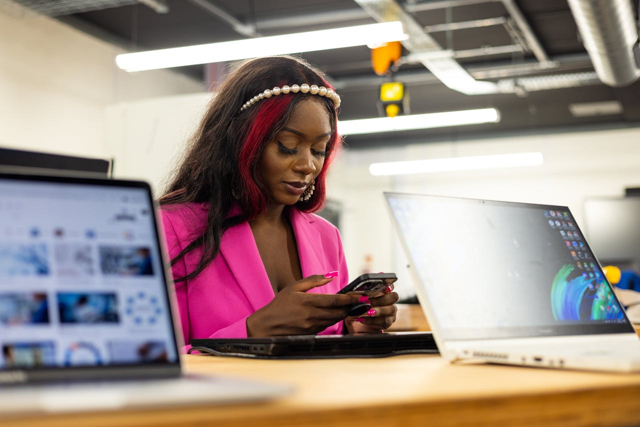A young woman in a vibrant pink suit using her smartphone at an office desk with laptops.
